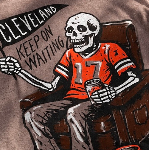 Close-up of a brown graphic t-shirt featuring a cartoon skeleton wearing an orange Cleveland Browns football jersey with the number 17, sitting in a chair and holding a drink. The text 'CLEVELAND KEEP ON WAITING' is printed on the left side of the graphic.
