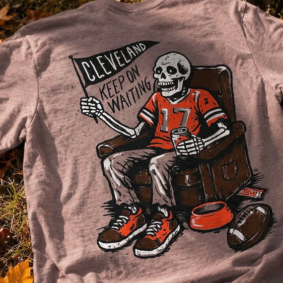 A brown graphic t-shirt laid flat on the ground outdoors, surrounded by autumn leaves and a tree trunk. The shirt features a cartoon skeleton wearing a Cleveland Browns football jersey with the number 17, sitting in a chair next to a football and a bowl, with a banner reading 'CLEVELAND KEEP ON WAITING'.